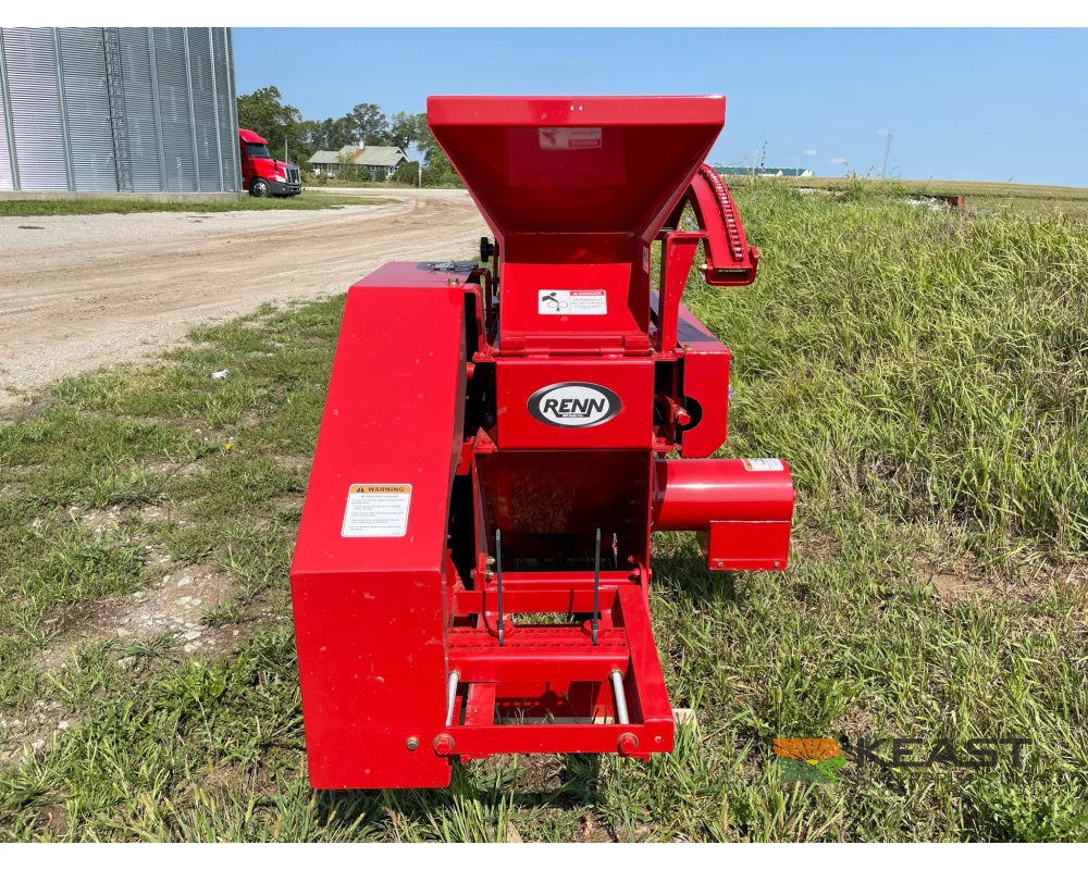 A red Renn 12 electric roller mill standing in grass at a dealer yard, seen from the discharge end with the feed hopper open above the rolls, the electric motor mounted on the right side and a grain bin and parked semi trailer on the gravel lot behind it