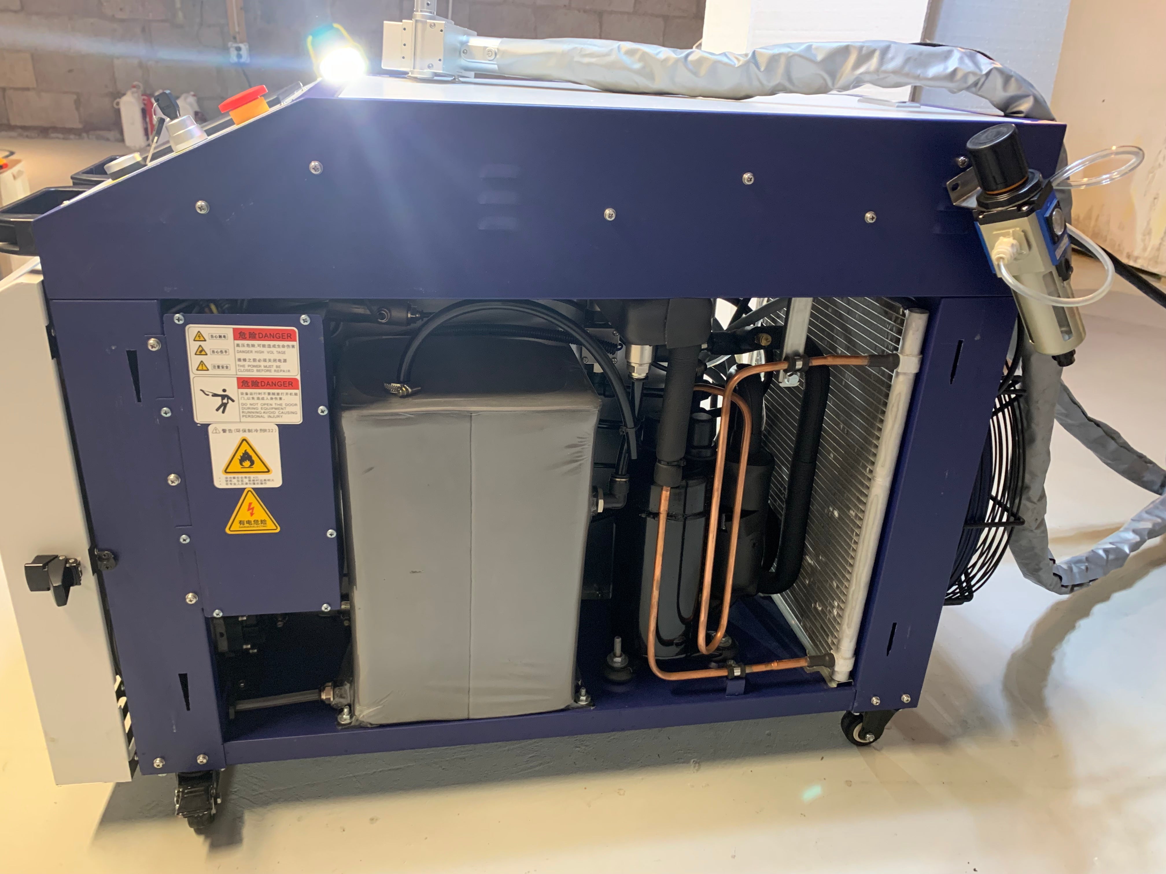 A blue fiber laser cleaner cabinet photographed in a block-walled shop with one side panel removed, showing the built-in water chiller's condenser coil, copper lines and fan, the insulated fiber conduit coiled across the top and laser warning labels on the frame